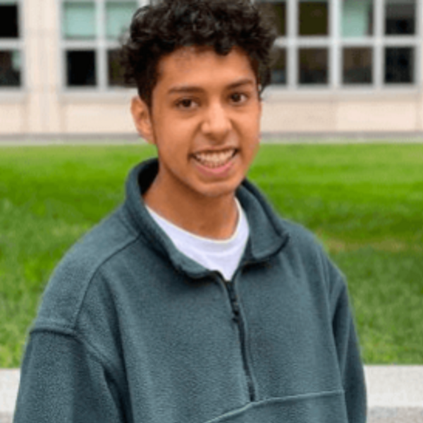 Hadin Sayed's headshot wearing a dark green fleece pullover. He has short brown curly hair and smiles in front of a grassy area and a building with large windows.