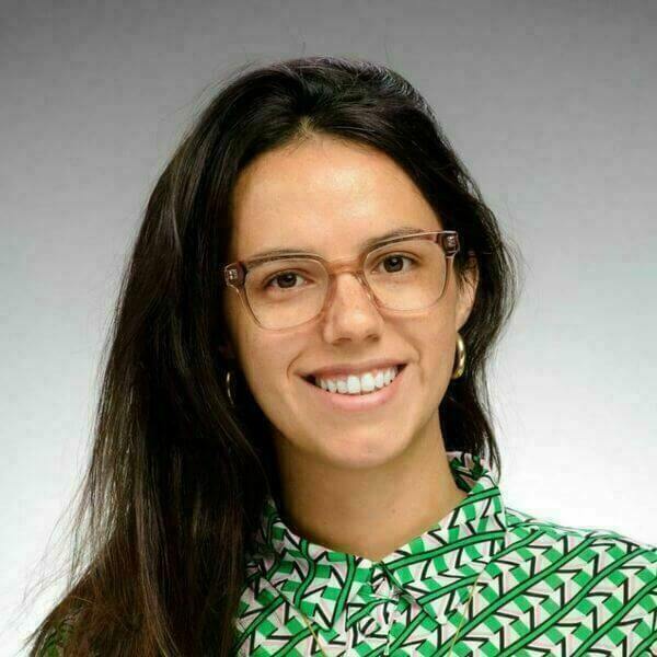 Headshot of Vicky Barone with long brown hair, wearing light-brown framed glasses and a green patterned shirt against a gray background. She is smiling at the camera.