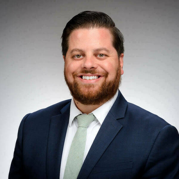 Headshot of Joe Waddington smiling against a gray background. He wears a navy suit jacket, white collared shirt, and sage green tie. He has short brown hair, a full reddish-brown beard, and is smiling broadly.