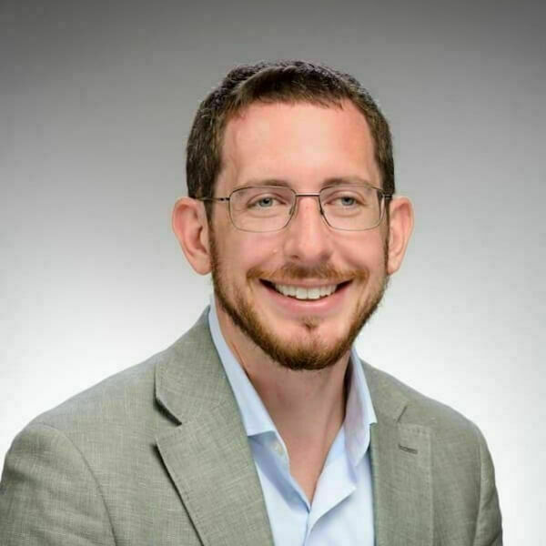 Headshot of Ryan Carpenter with short brown hair, glasses, and a beard. He wears a light gray blazer and light blue collared shirt. He smiles at the camera against a plain gray backdrop.