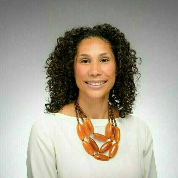 Headshot of Anna Haskins with curly brown hair, wearing a white shirt and a multi-strand necklace with oblong amber-colored beads. She is smiling at the camera against a gray background.