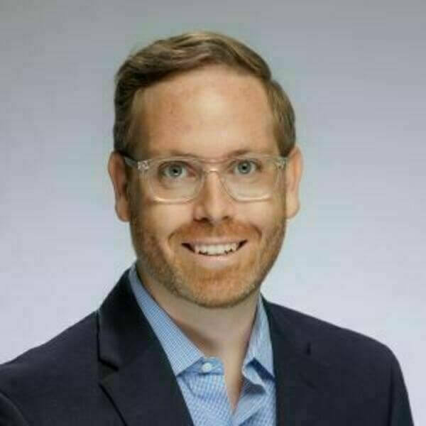 Headshot of Patrick Turner with light brown hair, clear-framed glasses, and a reddish-brown beard. He is wearing a dark blazer and a light blue button-down shirt. He is smiling against a plain gray background.