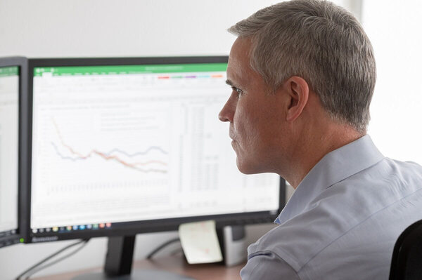 A man in a light blue, collared shirt sits and looks seriously at two computer monitors displaying line graphs. The graphs show a downward trend.