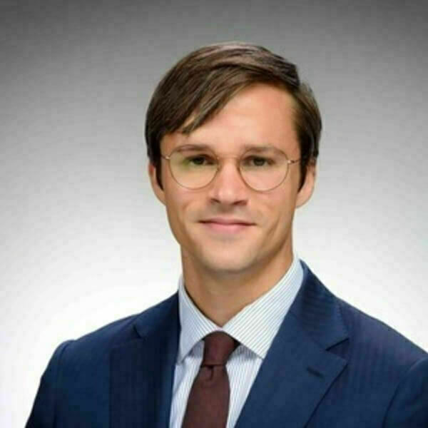 Headshot of John Tebes with short brown hair, wearing round eyeglasses, a dark blue suit, white pinstriped shirt, and a maroon tie.