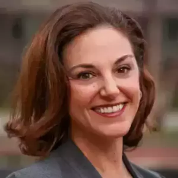 Melissa Kearney with brown hair, wearing a gray blazer and a pendant necklace. The outdoor background is softly blurred.