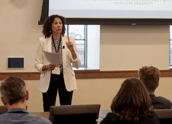 A woman, Anna Haskins, in a white blazer speaks, holding papers and gesturing with a raised index finger.