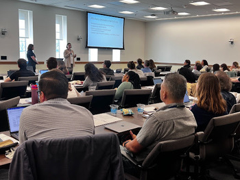 A woman speaks to a seated audience in a brightly lit room. A screen behind her reads 'Why Family Complexity is Hard to Study'.