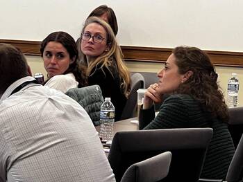 Three women at a meeting. Dark-haired woman looks back concerned. Blonde woman with glasses looks serious. Woman in green sweater looks thoughtful.