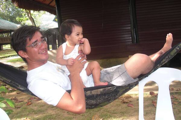 Smiling man in glasses holds a happy baby with fingers in mouth, both sitting in a patterned hammock outdoors.