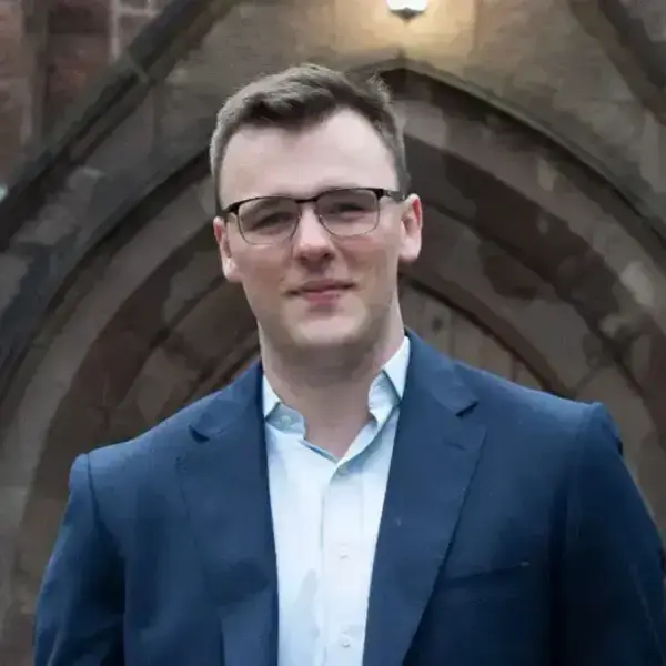 Young man with dark hair, glasses, wearing a navy blazer and light blue shirt, smiling slightly in front of a stone archway.