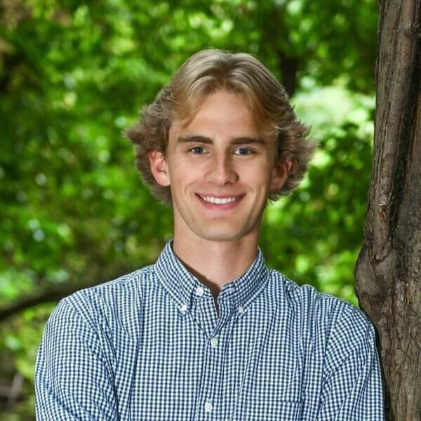 Young man with blond hair, blue eyes, and a white and blue checkered shirt smiles while leaning against a tree.